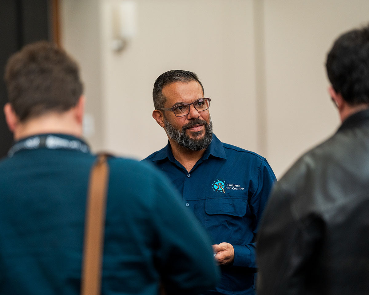 A close-up shot of a male industry professional with a beard and glasses in a blue collared work shirt speaking with colleagues during a networking break at the AEMEE conference.