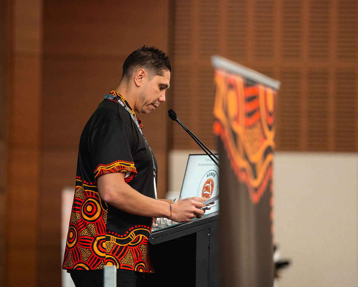 AEMEE CEO Jyi Lawton wearing a black and red traditional First Nations artwork shirt while speaking at a podium during the 2024 AEMEE conference.