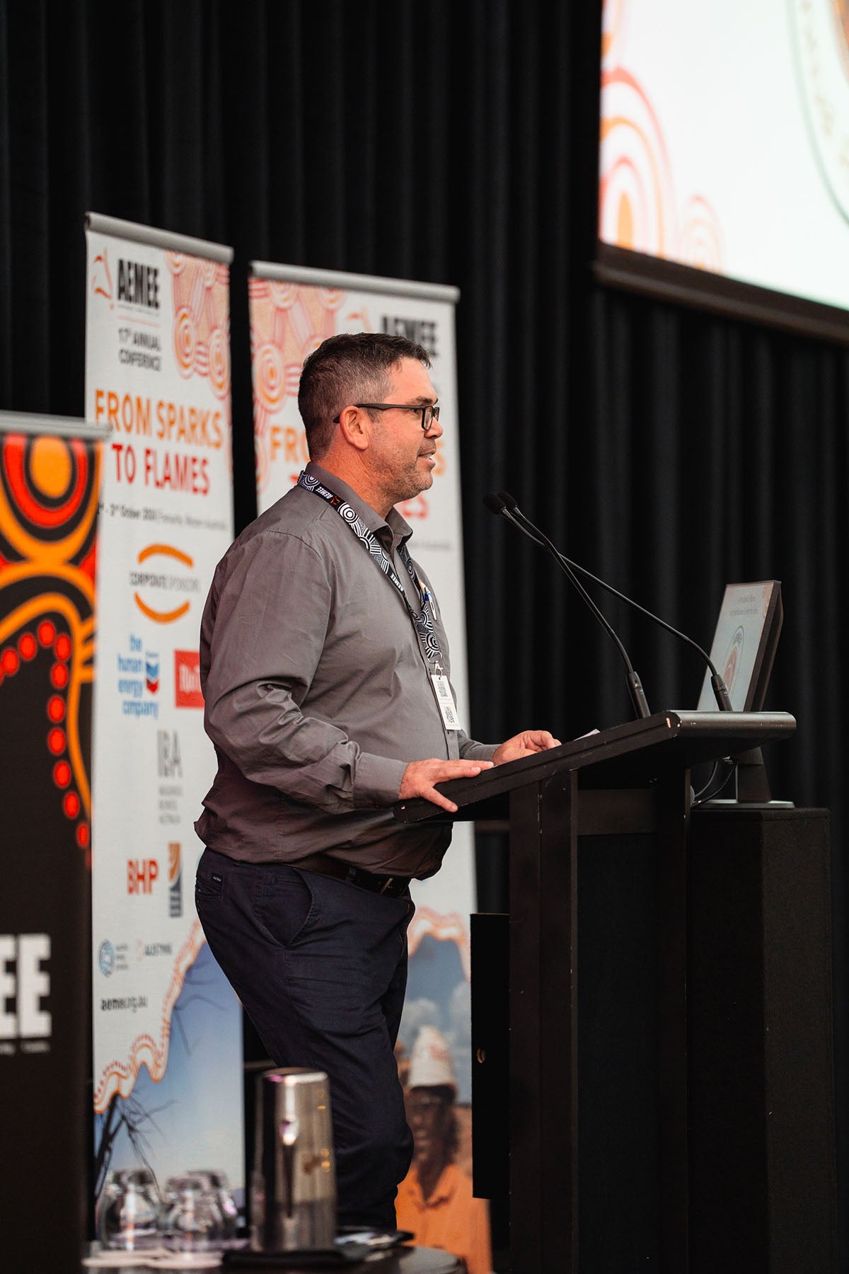 A male corporate speaker wearing glasses and a grey collared shirt presenting at the microphone during a resource panel breakout session at the AEMEE conference.
