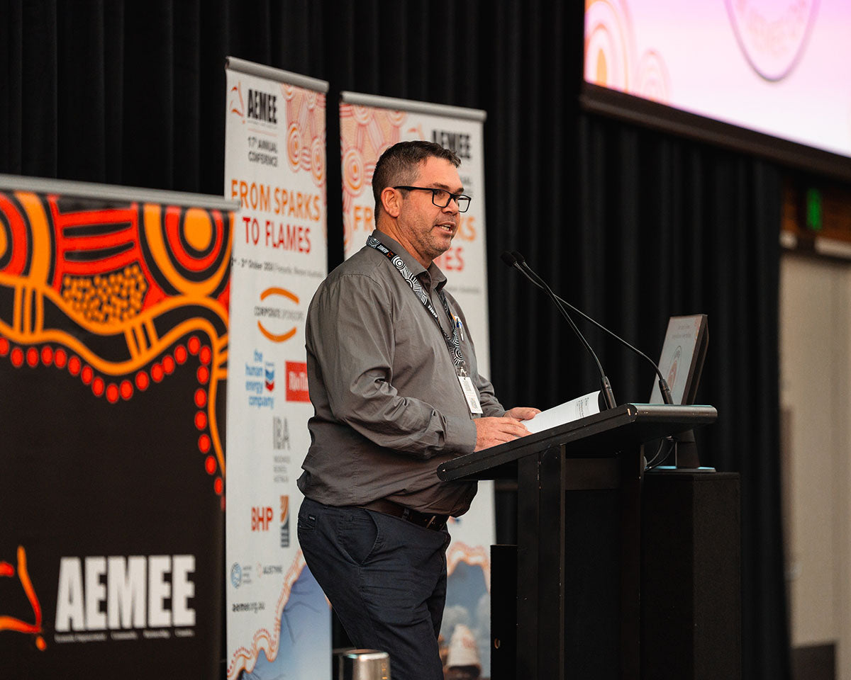 A male corporate speaker standing at a podium delivering an introductory address backdropped by a traditional First Nations media banner at the 17th Annual AEMEE conference.
