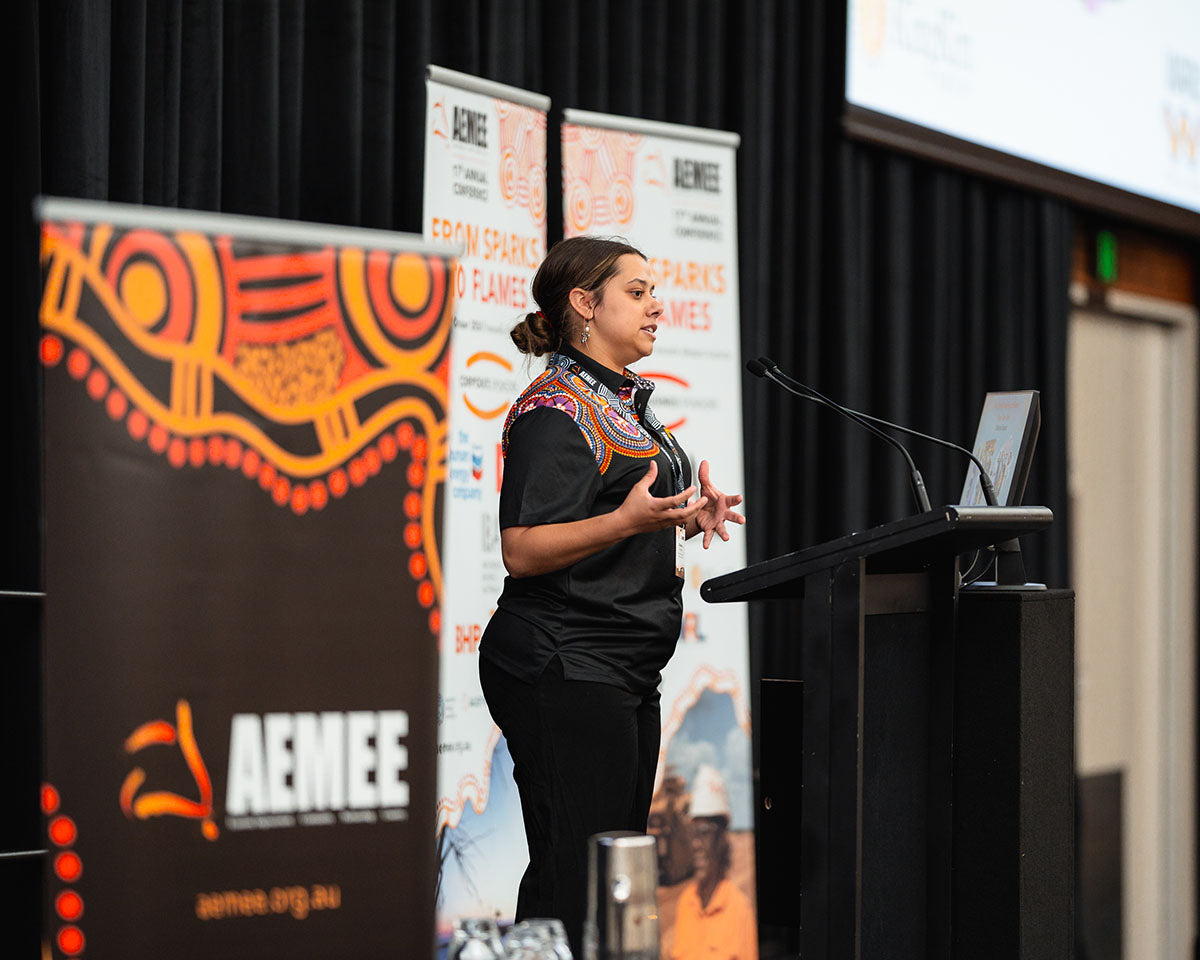 A female presenter wearing an intricate traditional artwork print collar standing at the stage podium during the 2024 Grounded Leadership session.