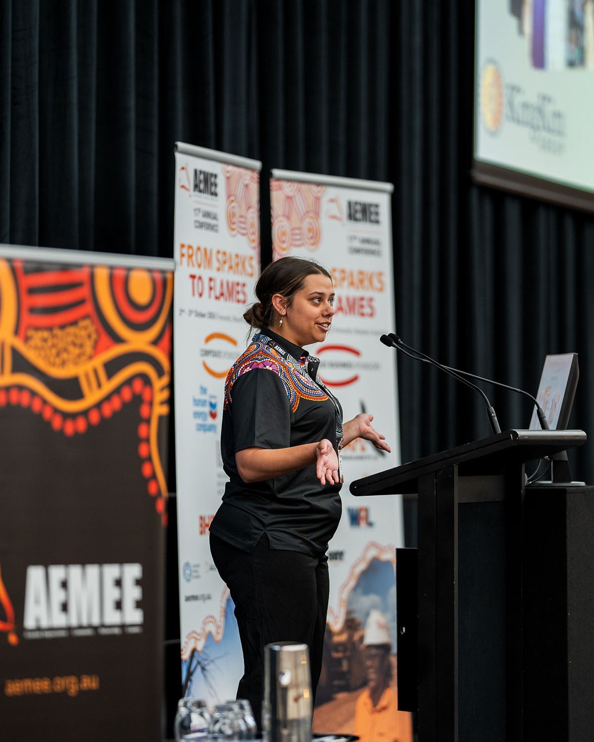 A female prAn overhead view of a fully packed presentation hall with seated industry delegates watching a keynote projector screen during a plenary panel at the AEMEE conference.esenter smiling while addressing the main auditorium crowd from the side podium at the annual national AEMEE conference.