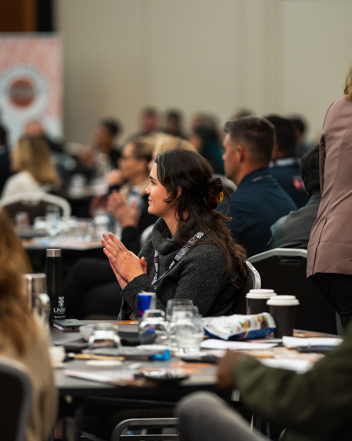 A side-profile view of an audie A presenter standing at the microphone beside a branded corporate media banner, addressing delegates within the Indigenous business resources sector.nce member applauding during an interactive panel discussion at the AEMEE conference.