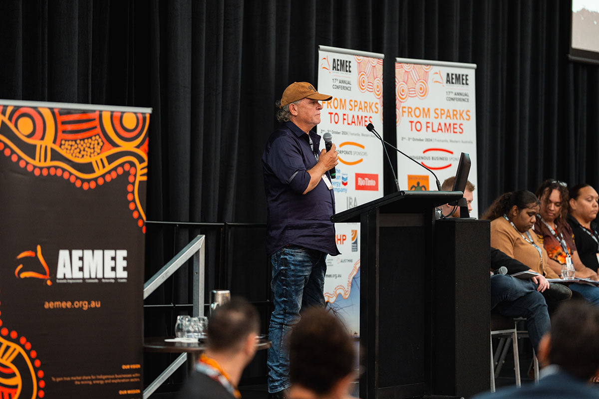 A presenter standing at the microphone beside a branded corporate media banner, addressing delegates within the Indigenous business resources sector.