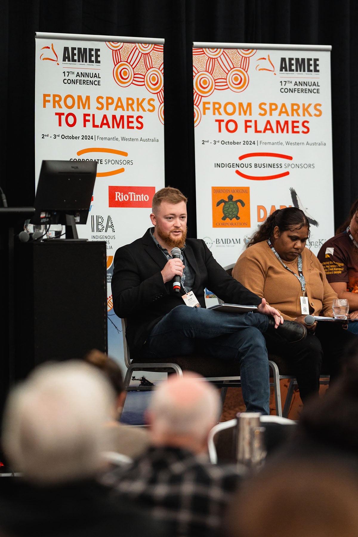 A male speaker addresses the audience into a microphone on stage backdropped by Rio Tinto and Baidam corporate sponsor banners at the AEMEE conference.