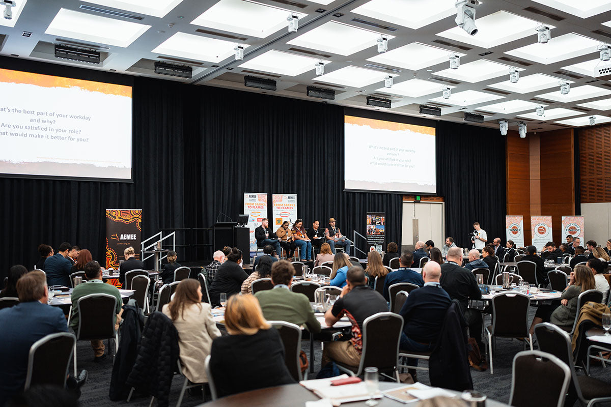 A wide perspective of a packed auditorium with seated audience tables watching a main-stage panel forum at the AEMEE conference.