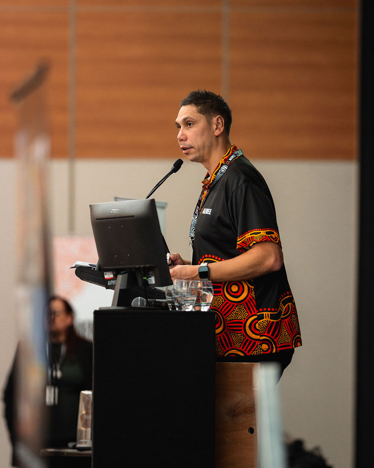 A close-up profile shot of AEMEE CEO Jyi Lawton addressing conference delegates and supply chain partners from the main stage podium.