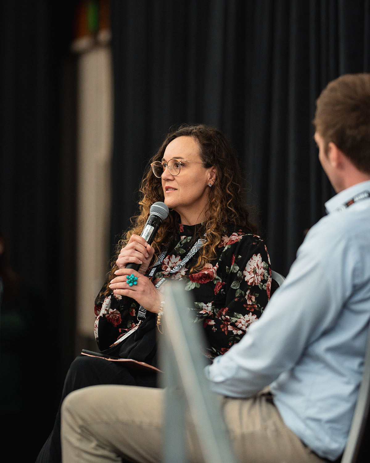 A female delegate wearing glasses and a floral blouse asks a question into a wireless microphone during a technical stream breakout session at the AEMEE conference.