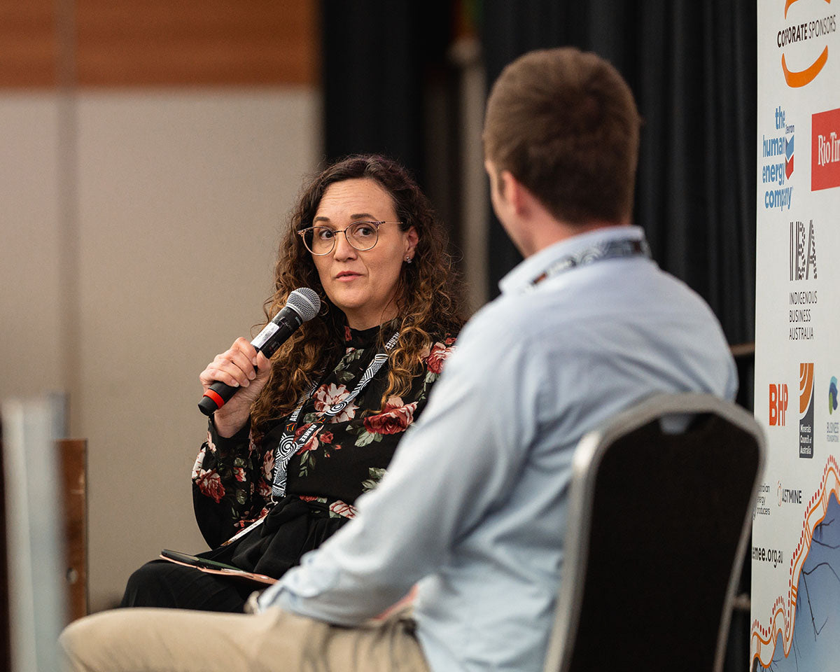 A female industry panelist wearing a floral blouse holding a microphone during an interactive stage discussion with a co-presenter at the annual AEMEE conference.