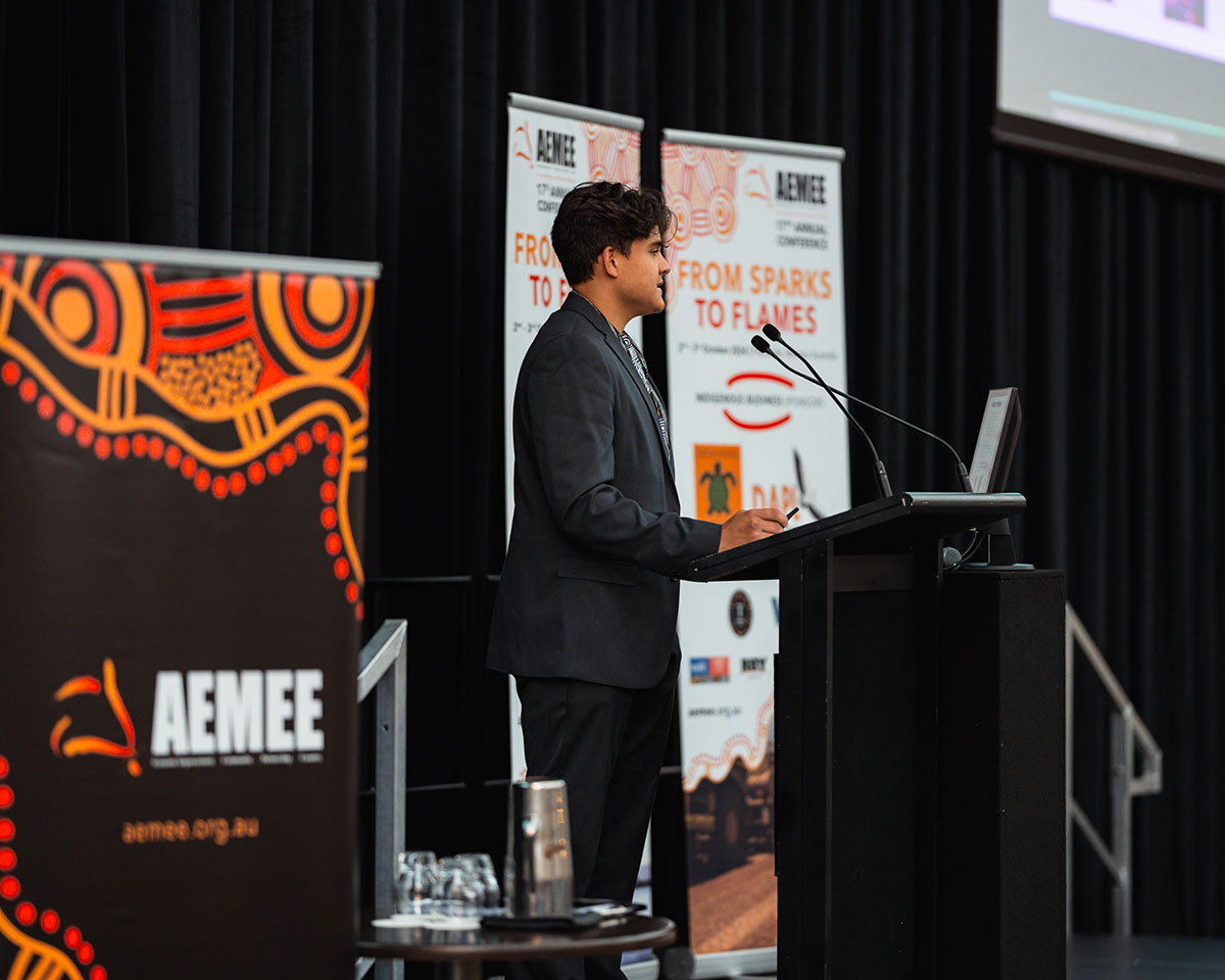 A young male industry representative in a dark blazer presenting from the stage podium beside a traditional First Nations graphic media banner at the AEMEE conference.