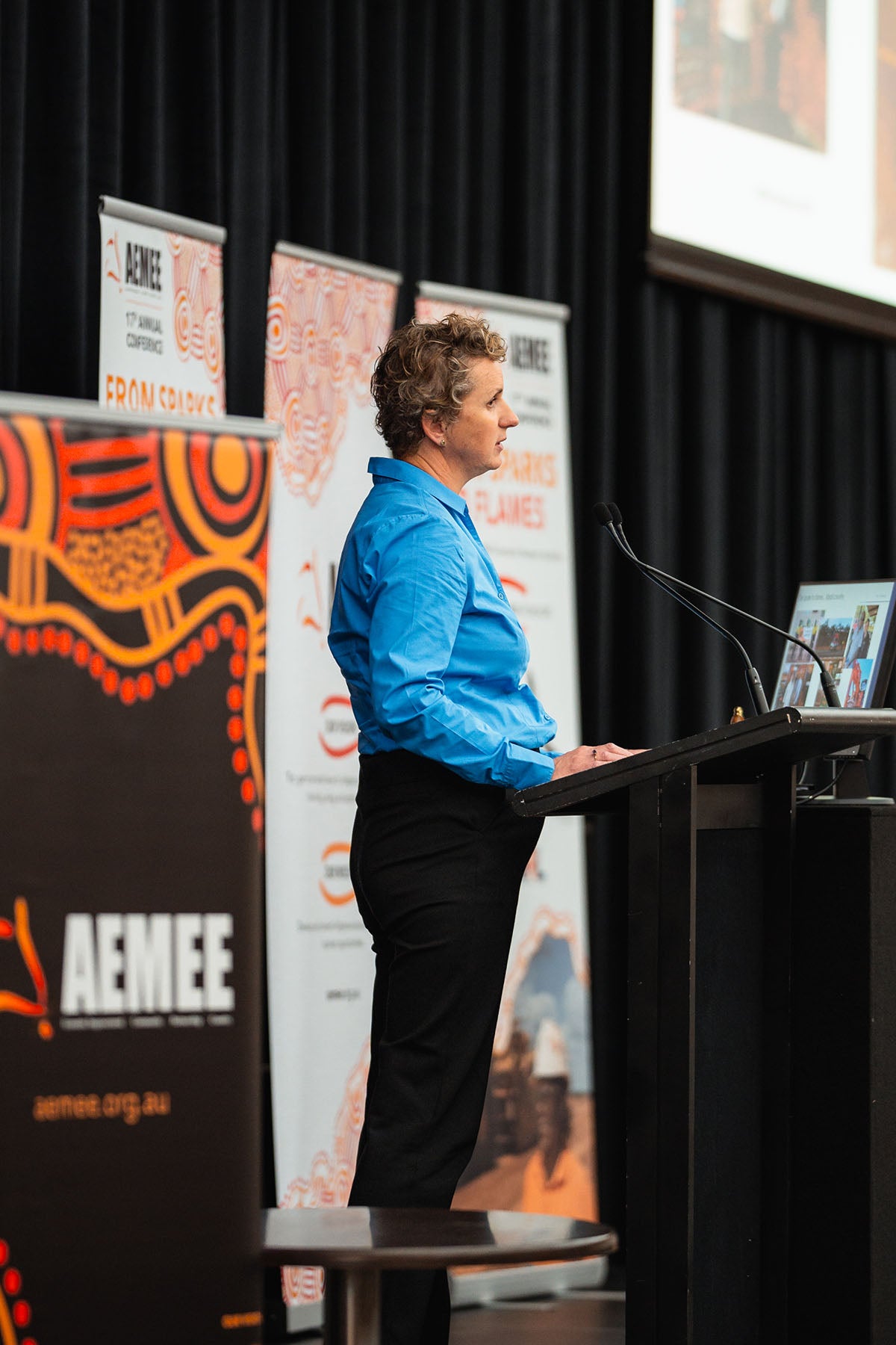 Susan O'Byrne of Business Foundations, wearing a sky blue shirt, delivering her presentation on sustainable financial independence for Aboriginal Corporations at the AEMEE conference podium.