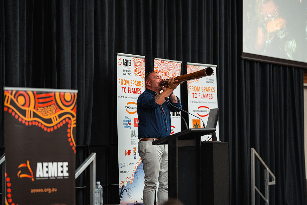 A cultural performer playing a traditional wooden yidaki at the microphone, backdropped by official "From Sparks to Flames" conference media banners.