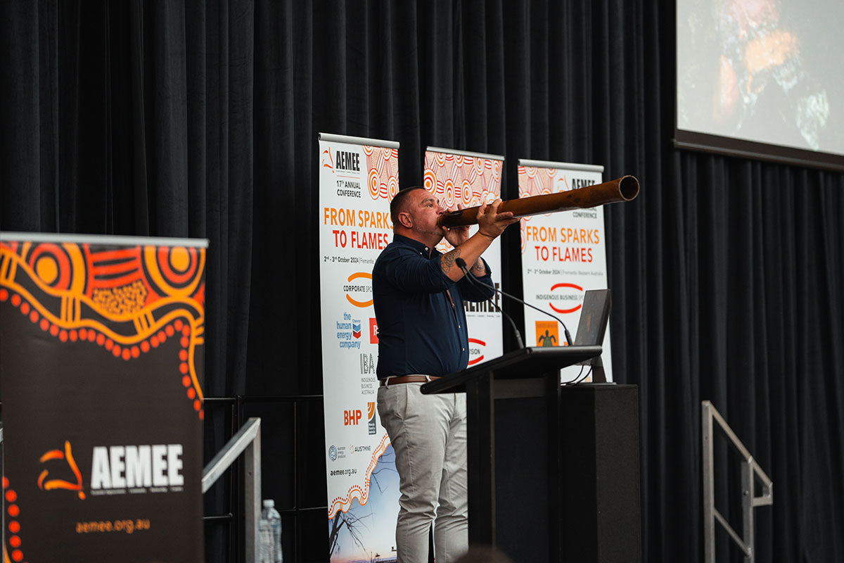 A male artist playing a traditional wooden didgeridoo on the main stage to mark the formal conclusion of the national AEMEE conference.