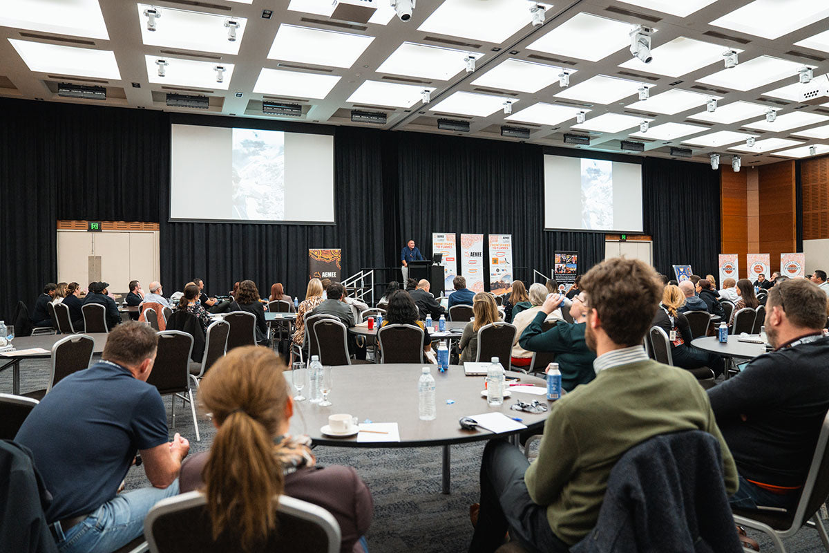 A wide view from the back of the auditorium showing seated delegates at round tables watching the active stage presentation at the annual national conference.