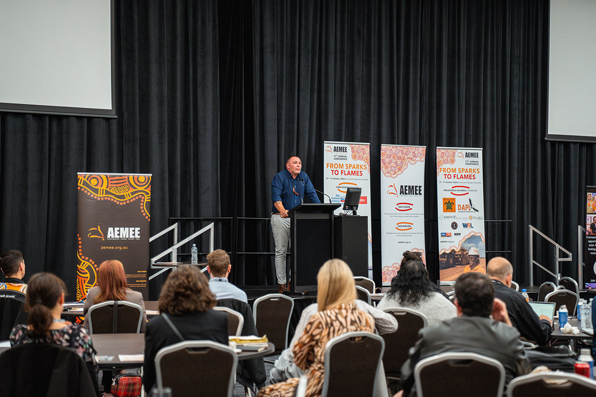A male speaker standing at the center stage podium addressing rows of seated industry delegates and business owners at the AEMEE conference.
