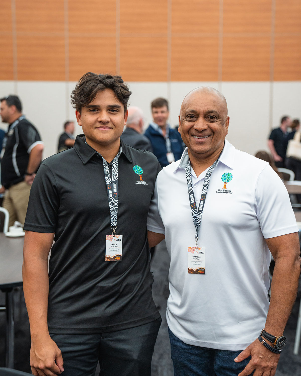 Two male corporate partners wearing conference lanyards and matching embroidered shirts smiling together inside the main hall during a networking block at the 17th Annual AEMEE conference.