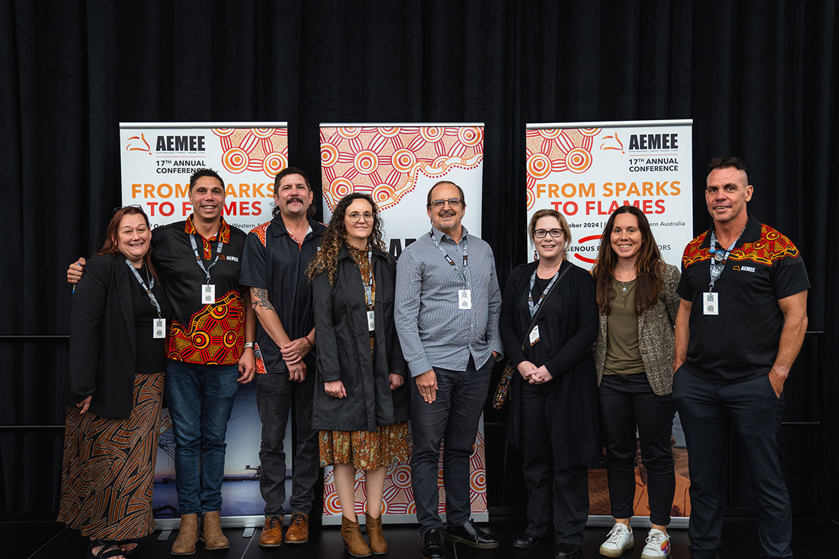 A group photo of nine corporate partners and event delegates, including AEMEE Chairperson Derek Flucker on the far right, standing together on stage in front of official "From Sparks to Flames" media banners.