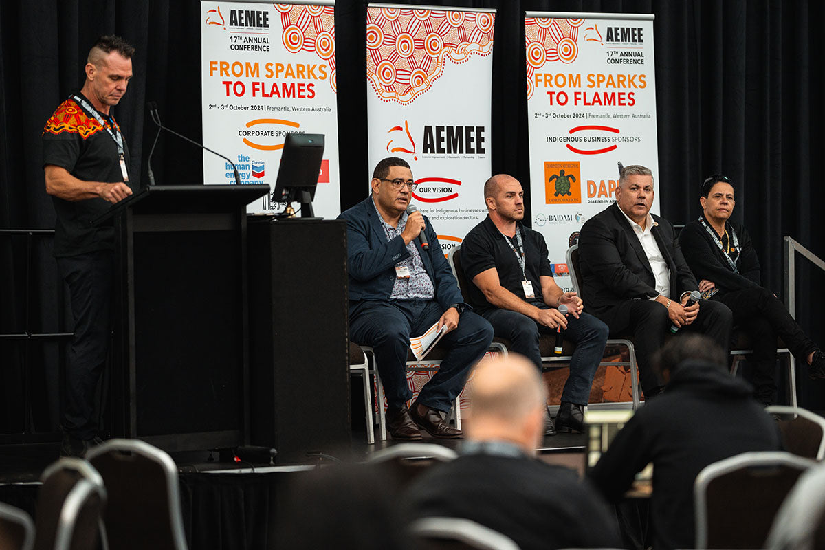 A presenter standing at the side podium introducing an interactive panel of four seated industry experts at the annual national AEMEE conference.