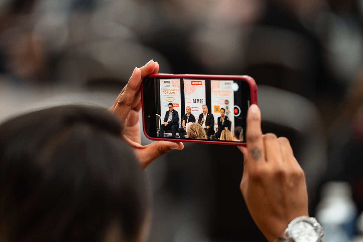 A close-up view of an audience member holding up a red smartphone to record a video of the active stage panel presentation at the AEMEE conference.