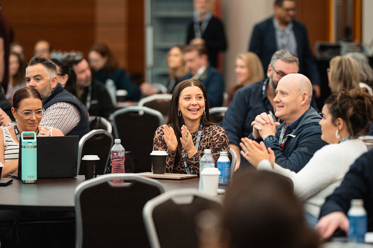 Corporate delegates and industry attendees smiling and applauding from their tables during an executive panel presentation at the AEMEE conference.