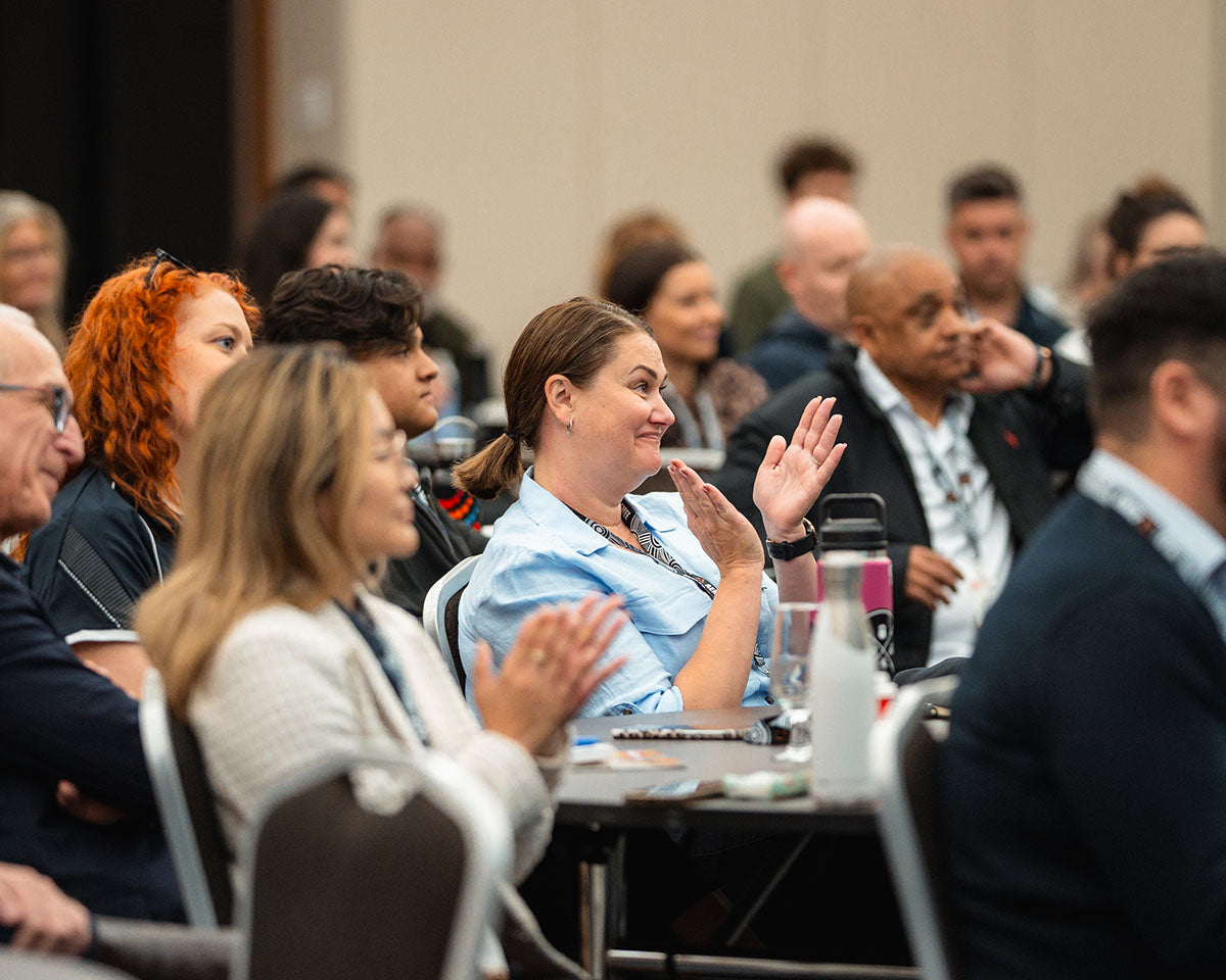 Corporate and community audience delegates smiling and clapping from their interactive tables during a plenary presentation at the annual AEMEE conference.