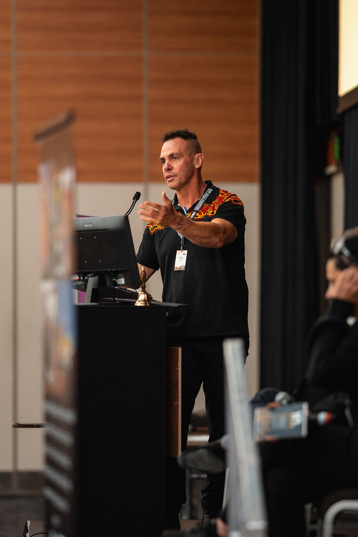 AEMEE Chairperson and RBY Projects Managing Director Derek Flucker delivering an executive address with an open-hand gesture from the stage podium.