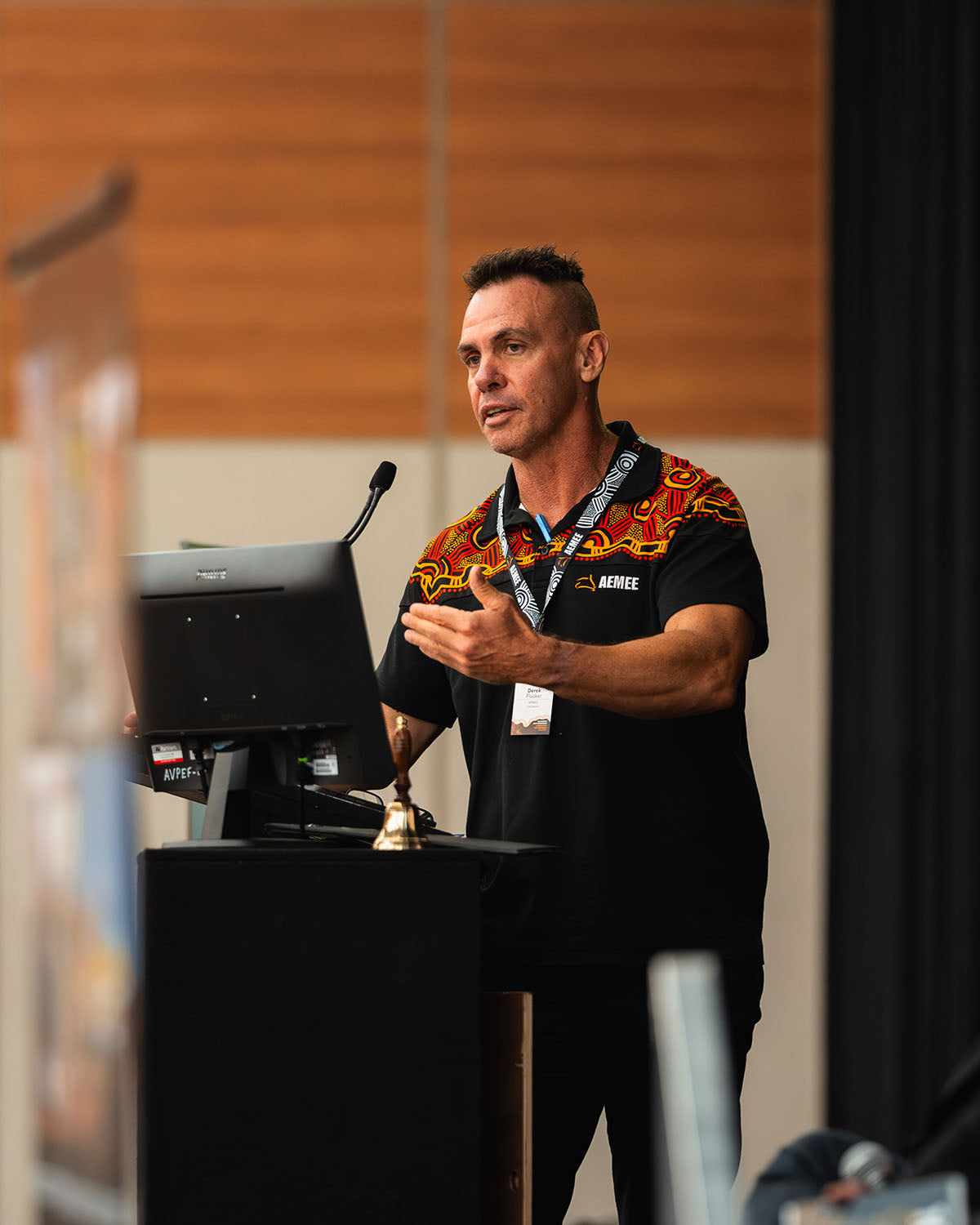 A close-up view of AEMEE Chairperson Derek Flucker speaking at the microphone, wearing a corporate shirt featuring traditional First Nations artwork designs.