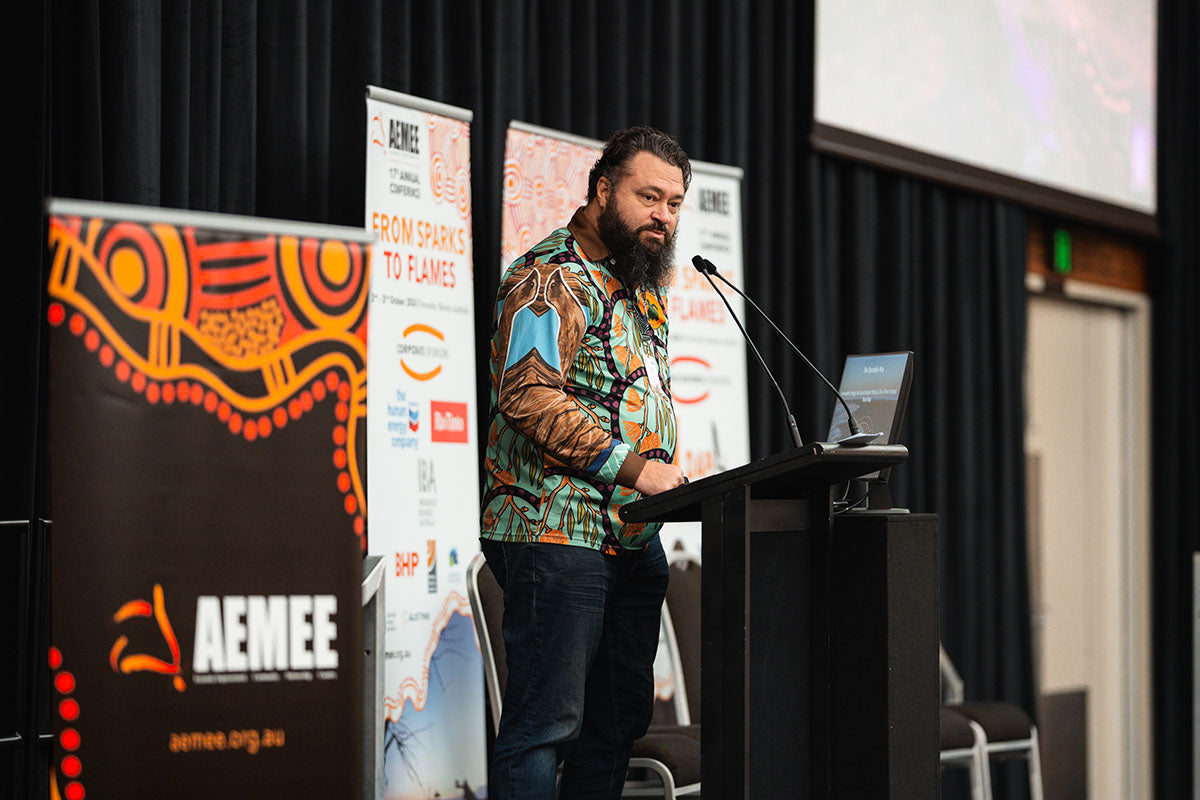 Nathan McIvor from Djarindjin, wearing a vibrant patterned artwork shirt, presenting "The Djarindjin Way" strategic road map from the podium at the annual AEMEE conference.