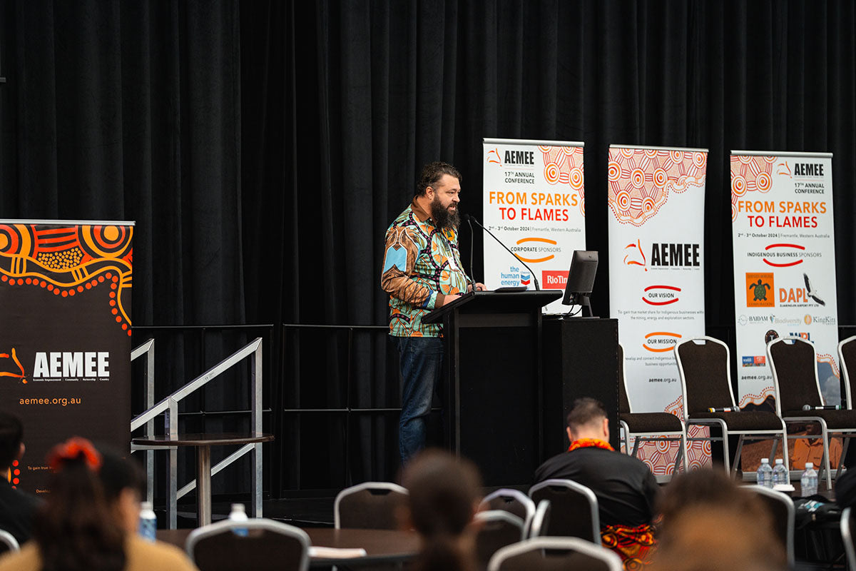 Presenter Nathan McIvor standing at the main stage podium under corporate media banners during his sustainable development session at the national AEMEE conference.