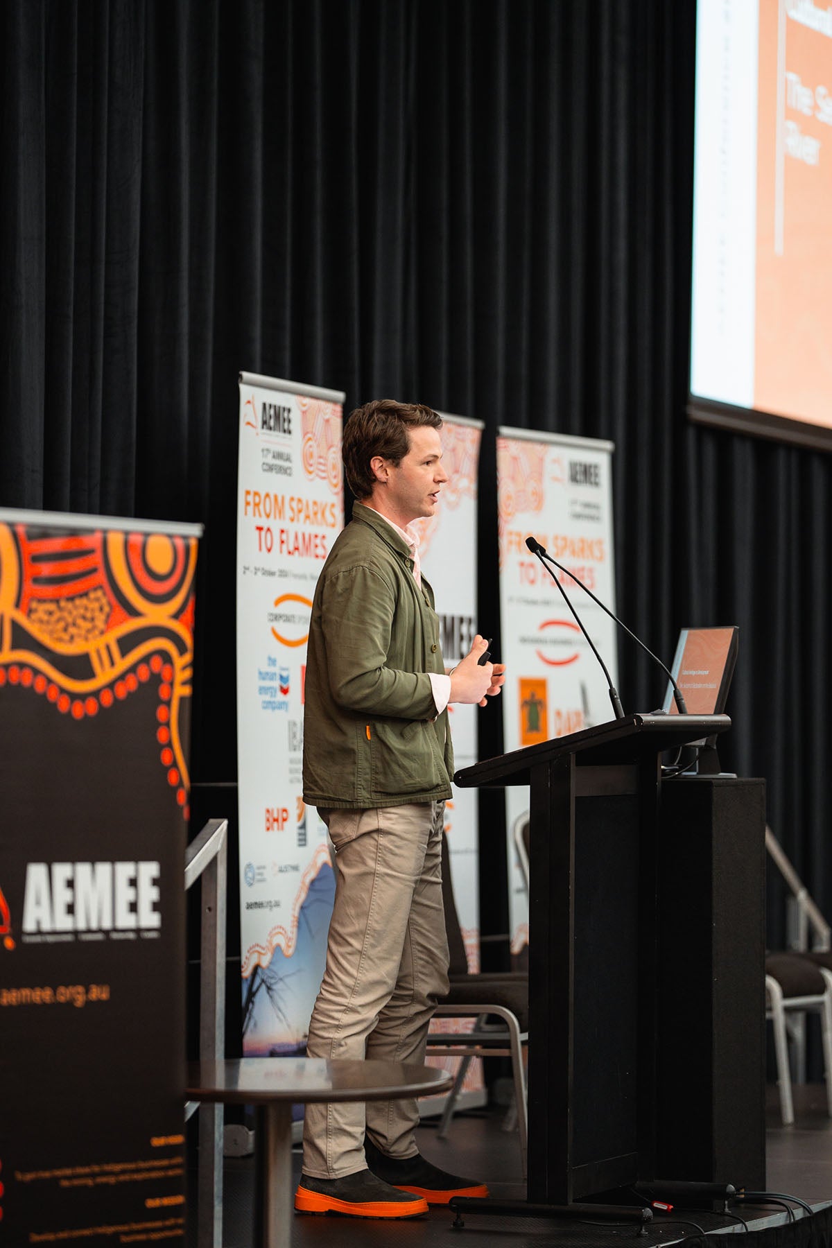 A male industry professional in a green jacket speaking at the podium alongside a "From Sparks to Flames" media banner at the annual national AEMEE conference.