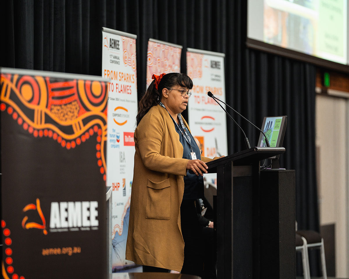 Denise Stan from Sherlock Contracting, wearing a long brown coat, addressing corporate delegates from the stage podium during a technical stream presentation.