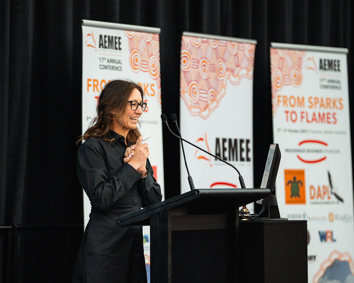 A female presenter wearing glasses and a black collared shirt smiling with hands clasped at the stage podium during the 17th Annual AEMEE conference.