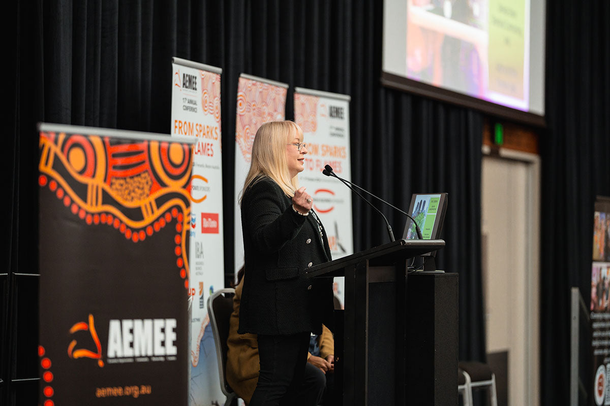 Sabina Schlink of Thomas Legal, a blonde presenter in a black blazer, gesturing while addressing the audience from the stage podium at the annual AEMEE conference.