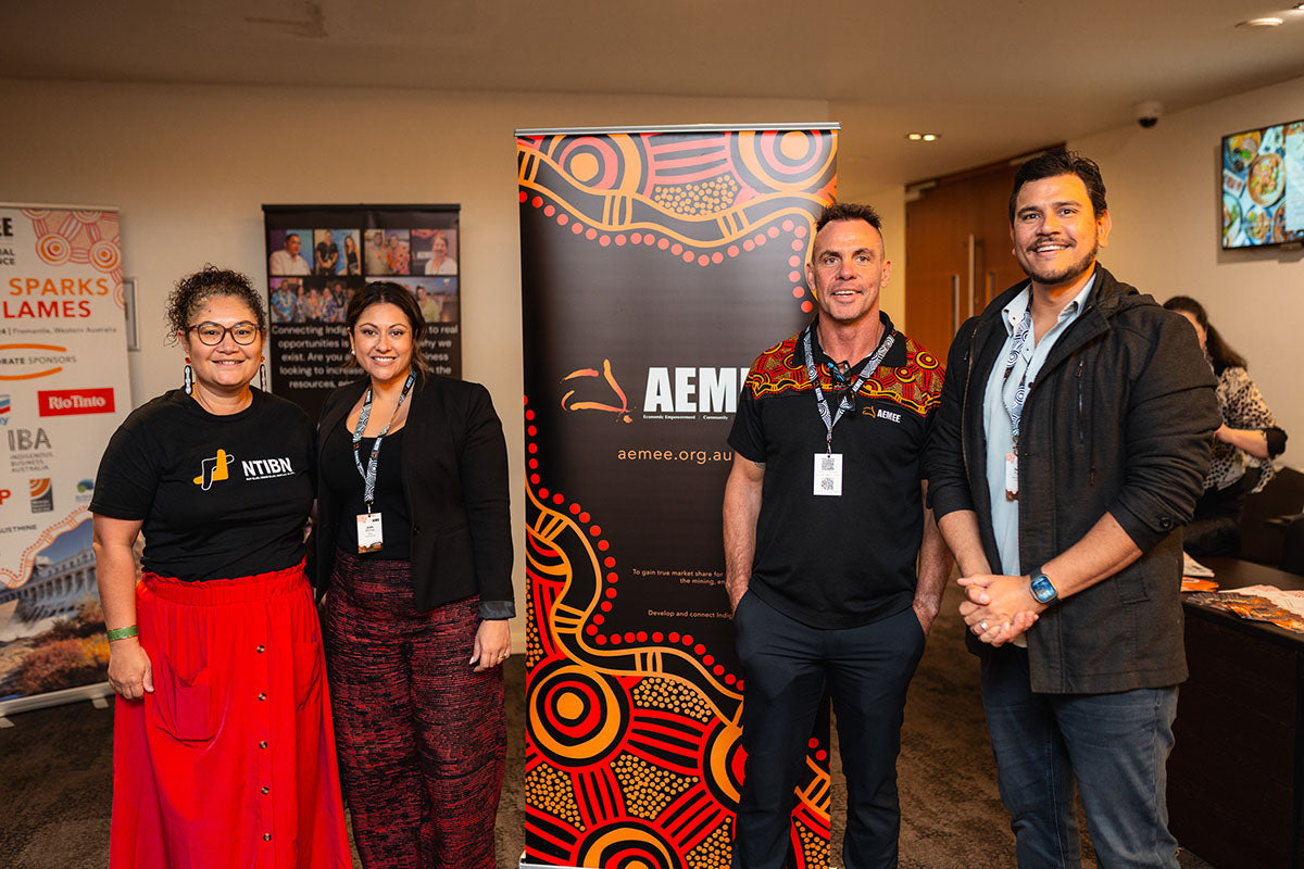 An NTIBN representative, AEMEE board member and DICE Australia Director Raymond Pratt, AEMEE Chairperson Derek Flucker, and an industry delegate standing together smiling in front of official promotional media banners at the 2024 conference.