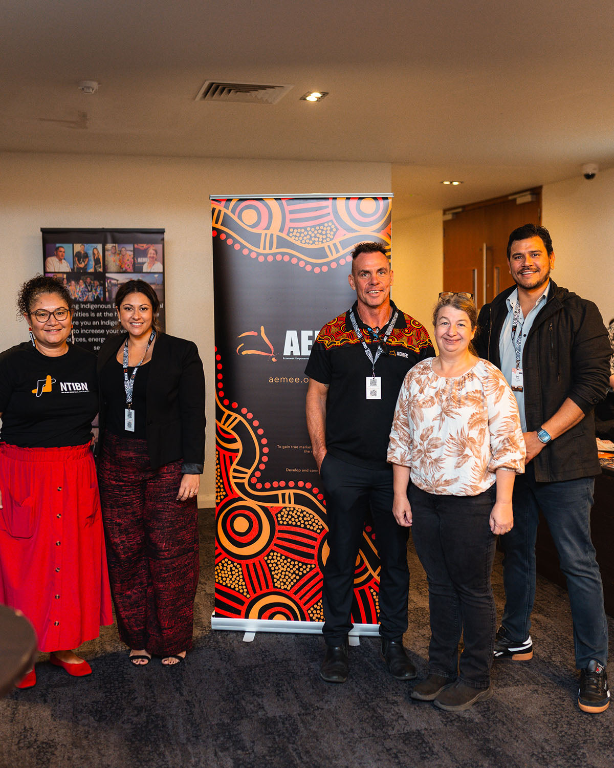 An NTIBN representative, AEMEE board member and DICE Australia Director Raymond Pratt, AEMEE Chairperson Derek Flucker, and an industry delegate standing together smiling in front of official promotional media banners.