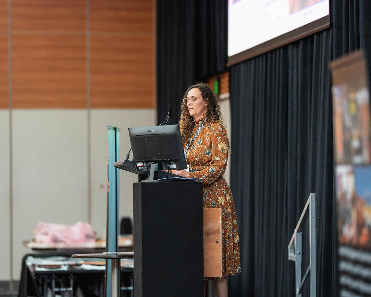 A female industry representative wearing a patterned brown dress delivering an address from the stage podium during a breakout session at the AEMEE conference.