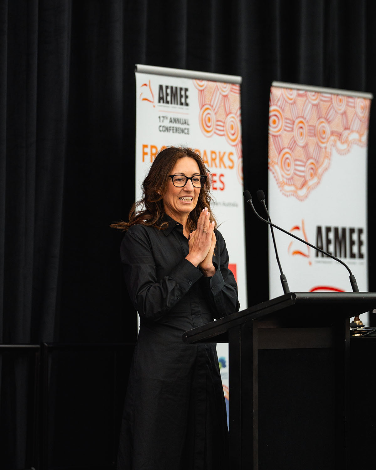 A female industry leader addressing conference delegates from the main stage podium, backdropped by official "From Sparks to Flames" media banners.