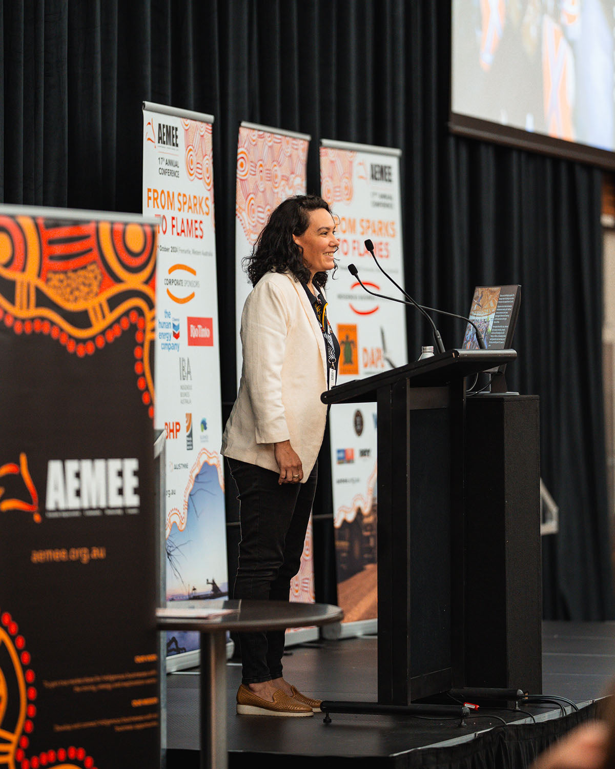 A female speaker wearing a white blazer presenting from the stage podium beside a "From Sparks to Flames" corporate sponsor banner.