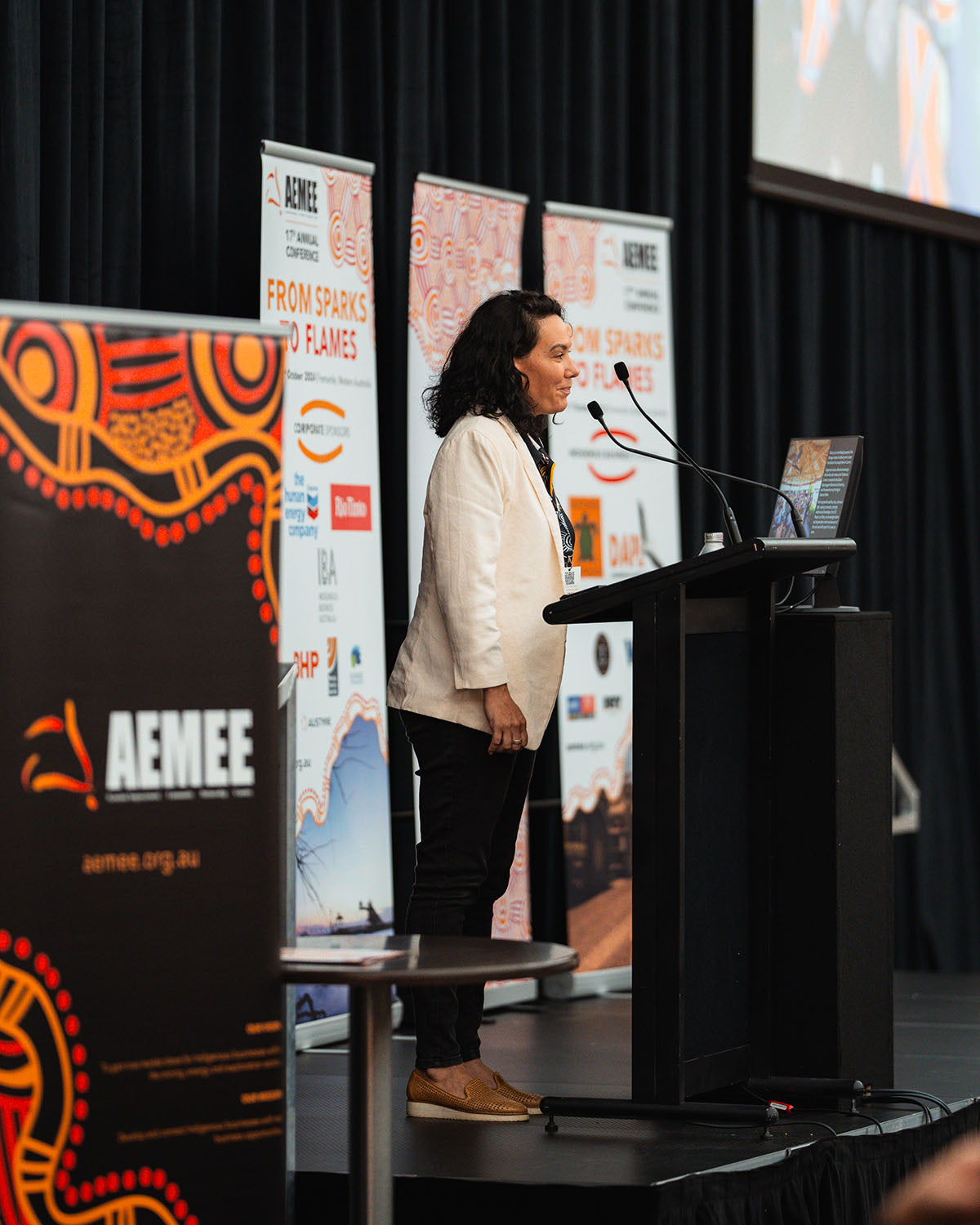 A female presenter in a white blazer speaking at the microphone during an interactive stream focused on commercial pathways at the national AEMEE conference.