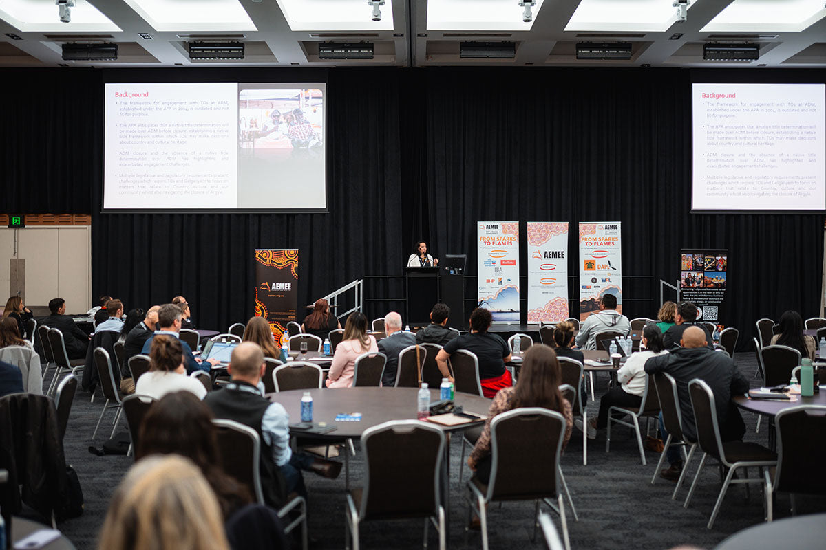 A wide view of a packed conference auditorium with rows of round tables filled with delegates watching an active stage presentation beneath large overhead projector screens.
