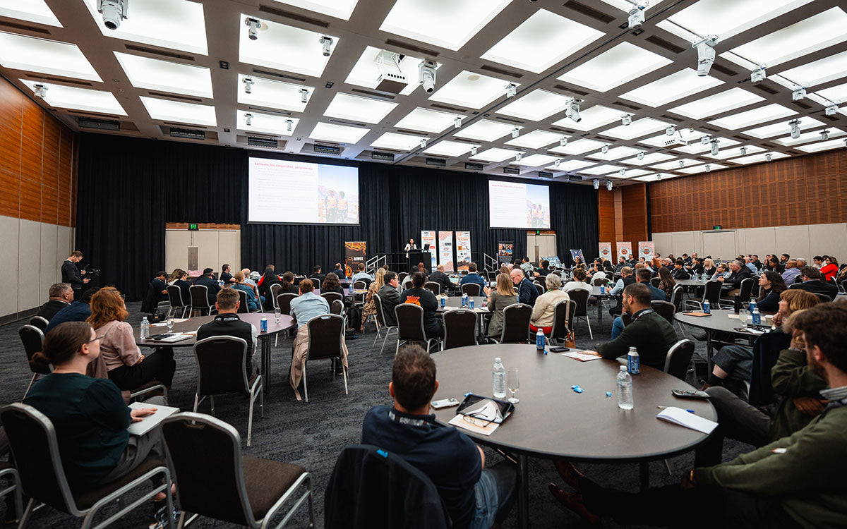 A wide view from the back of the conference auditorium showing delegates seated at round tables facing the presentation stage during a plenary session at the AEMEE conference.
