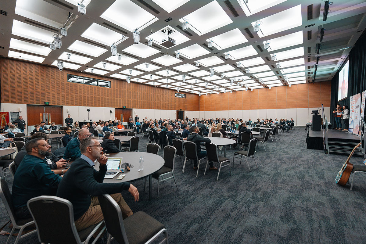Seated business professionals and delegates looking toward the stage beneath a large overhead projector display at the national AEMEE conference.