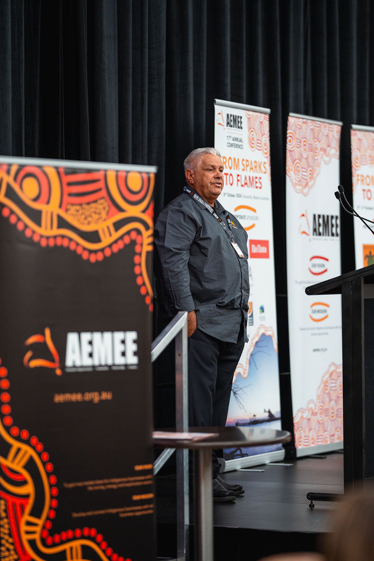A male industry representative gesturing with an open hand while speaking next to a podium backdropped by traditional First Nations art media banners at the AEMEE conference.