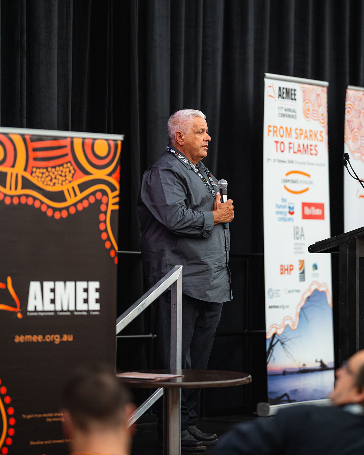 A male corporate speaker standing on stage beside a "From Sparks to Flames" banner during a presentation on strategic pathways within the Indigenous business resources sector.