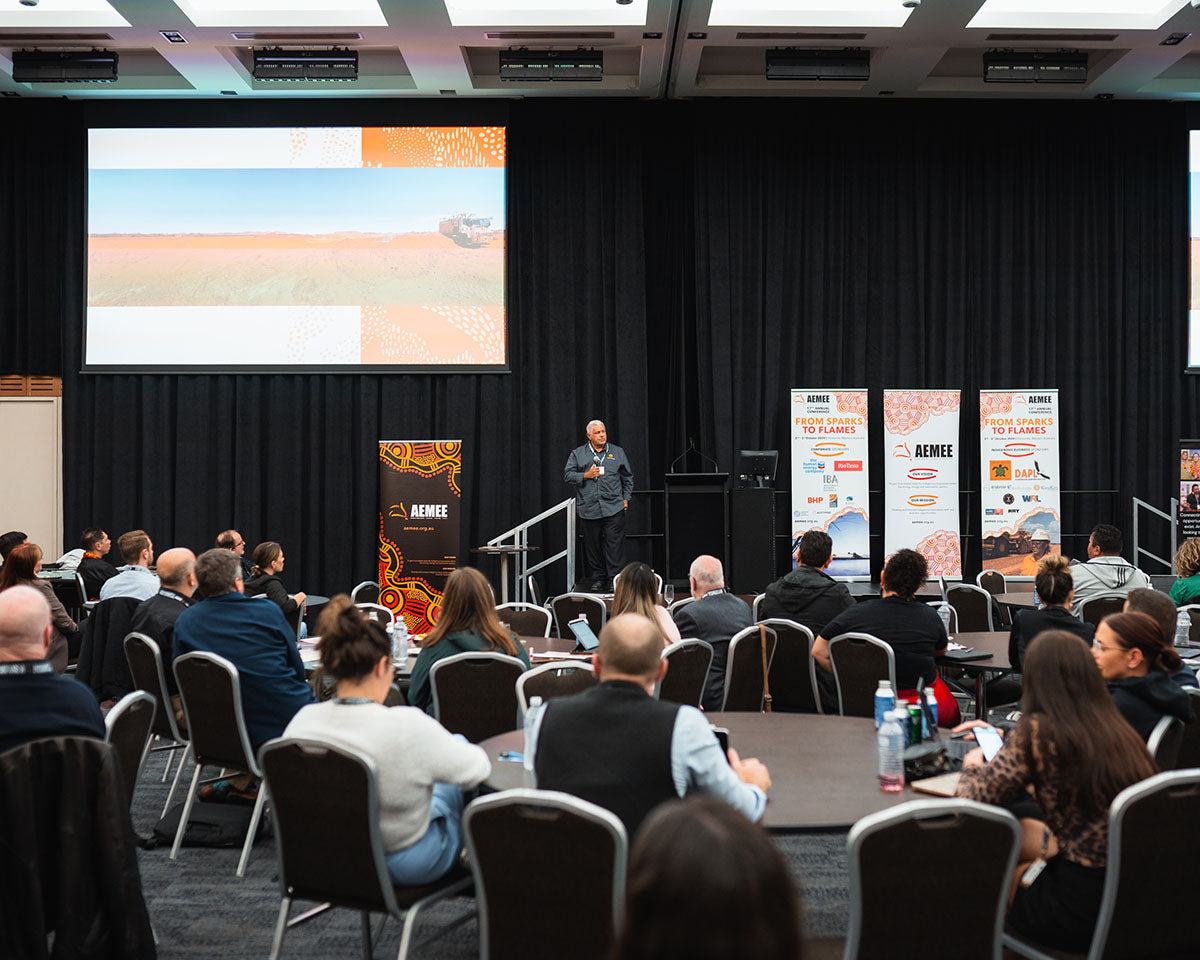 A presenter standing on stage speaking into a microphone to an auditorium of seated delegates during a charity auction stream at the annual AEMEE conference.