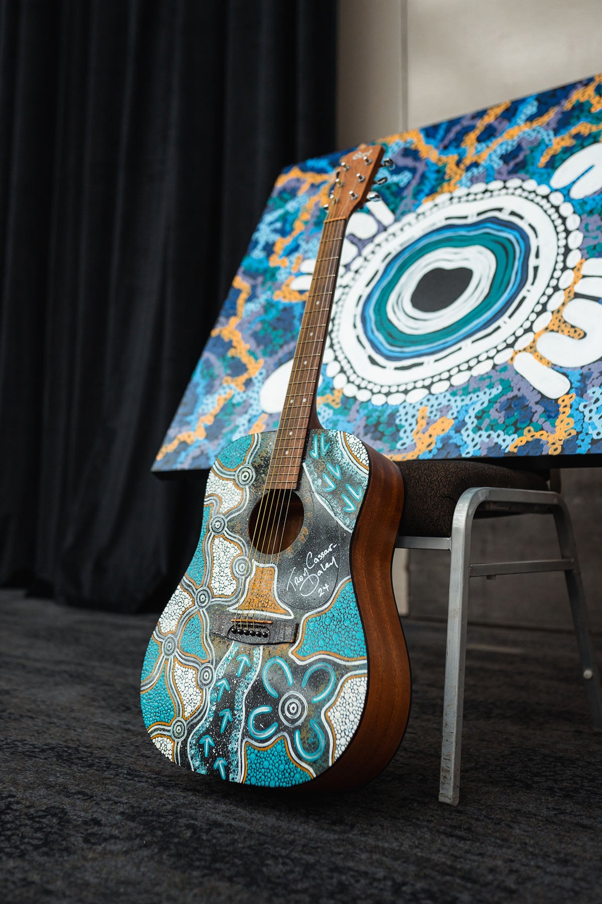 A custom-painted acoustic guitar standing in front of a matching large-scale canvas dot artwork during a fundraising showcase for the Indigenous business resources sector.