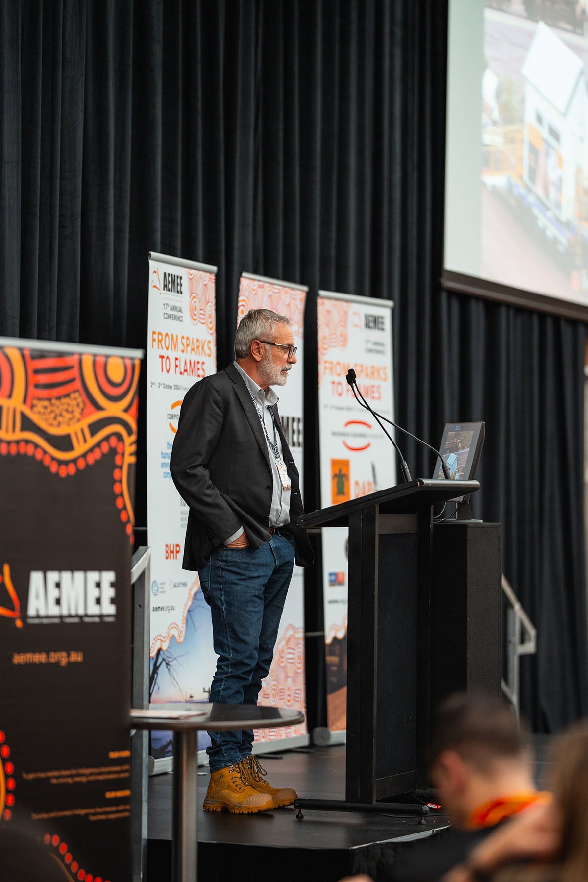 An industry representative with glasses and a gray beard speaking at the stage microphone, backdropped by official "From Sparks to Flames" media banners.