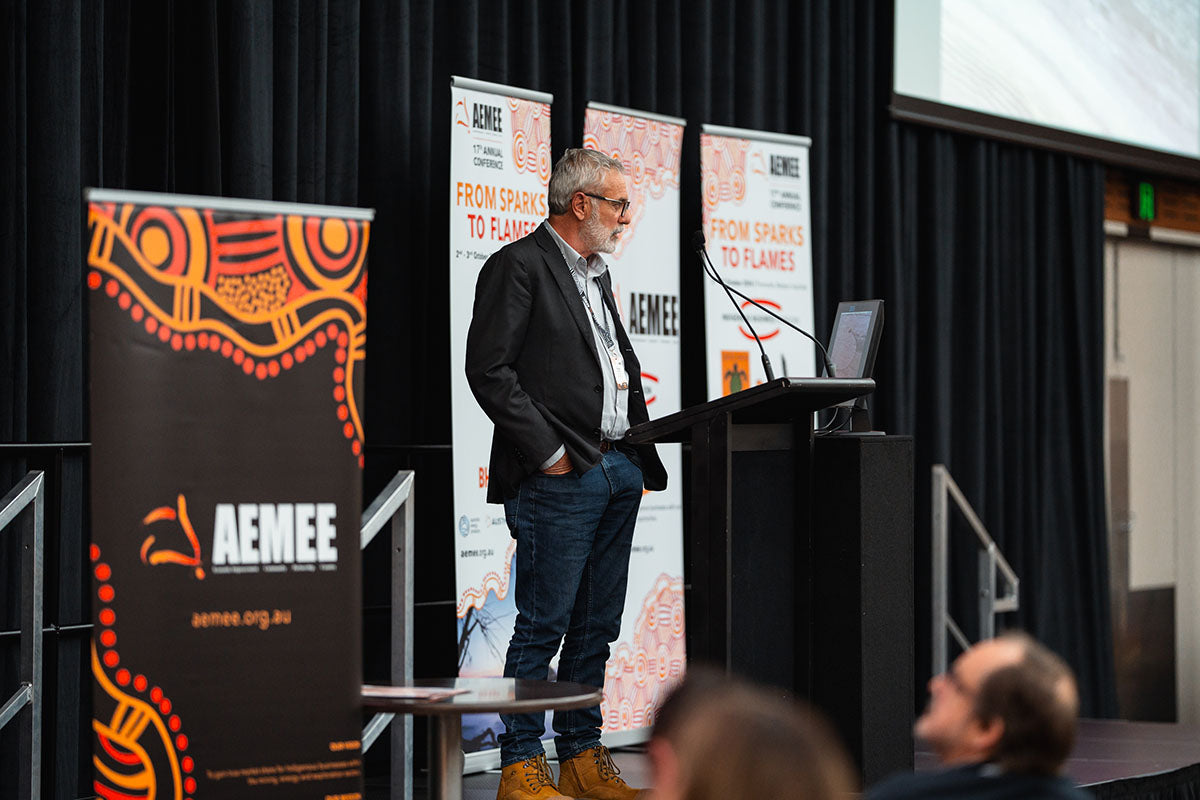 Speaker standing near the stage podium and speaking into the microphone during his presentation, with official “From Sparks to Flames” media banners in the background.