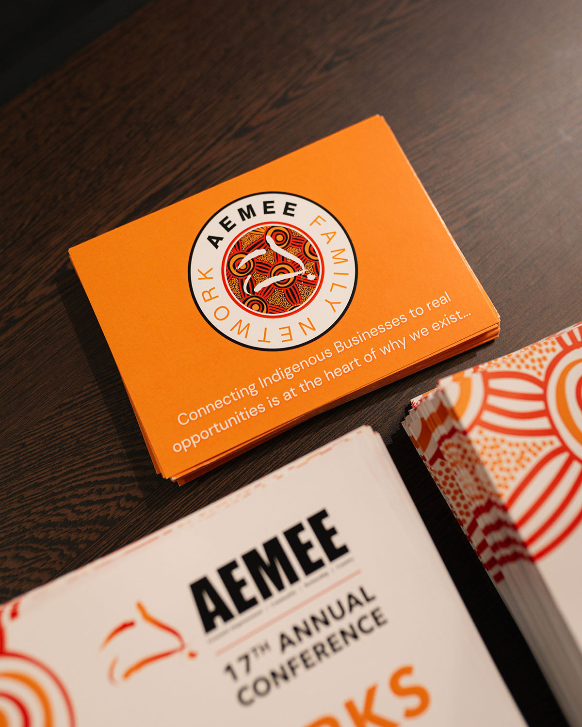A close-up shot of a stack of orange AEMEE Family Network marketing cards featuring the slogan "Connecting Indigenous Businesses to real opportunities is at the heart of why we exist."