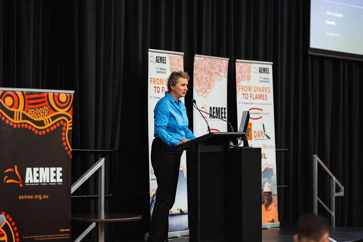 A female speaker in a blue shirt addressing the auditorium crowd from the side podium under corporate partner banners at the national AEMEE conference.